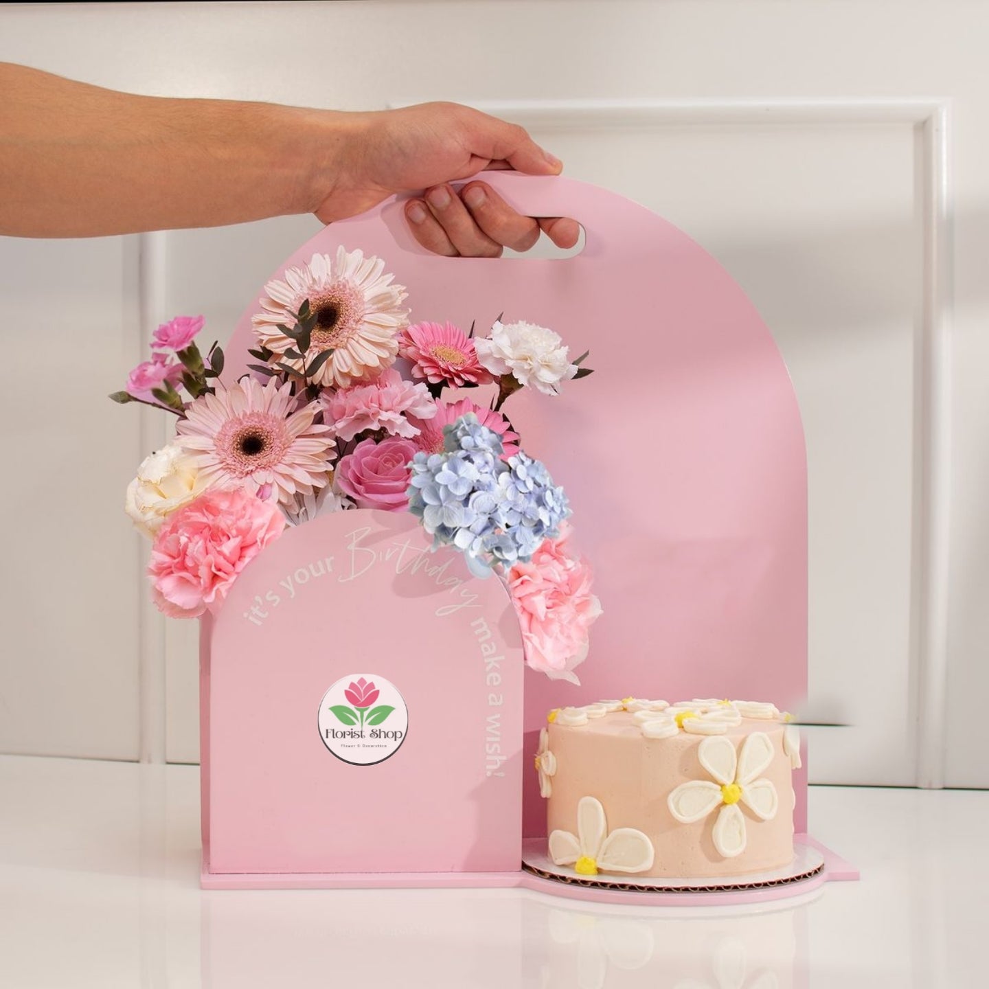 Pink floral arrangement in a box with a cake on a white surface, featuring a visible brand logo.