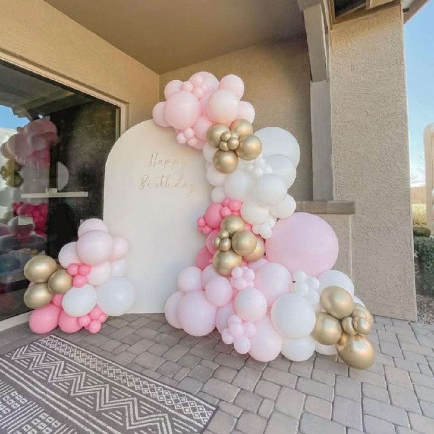 Balloon arch with pink, gold, and white balloons in front of a house.