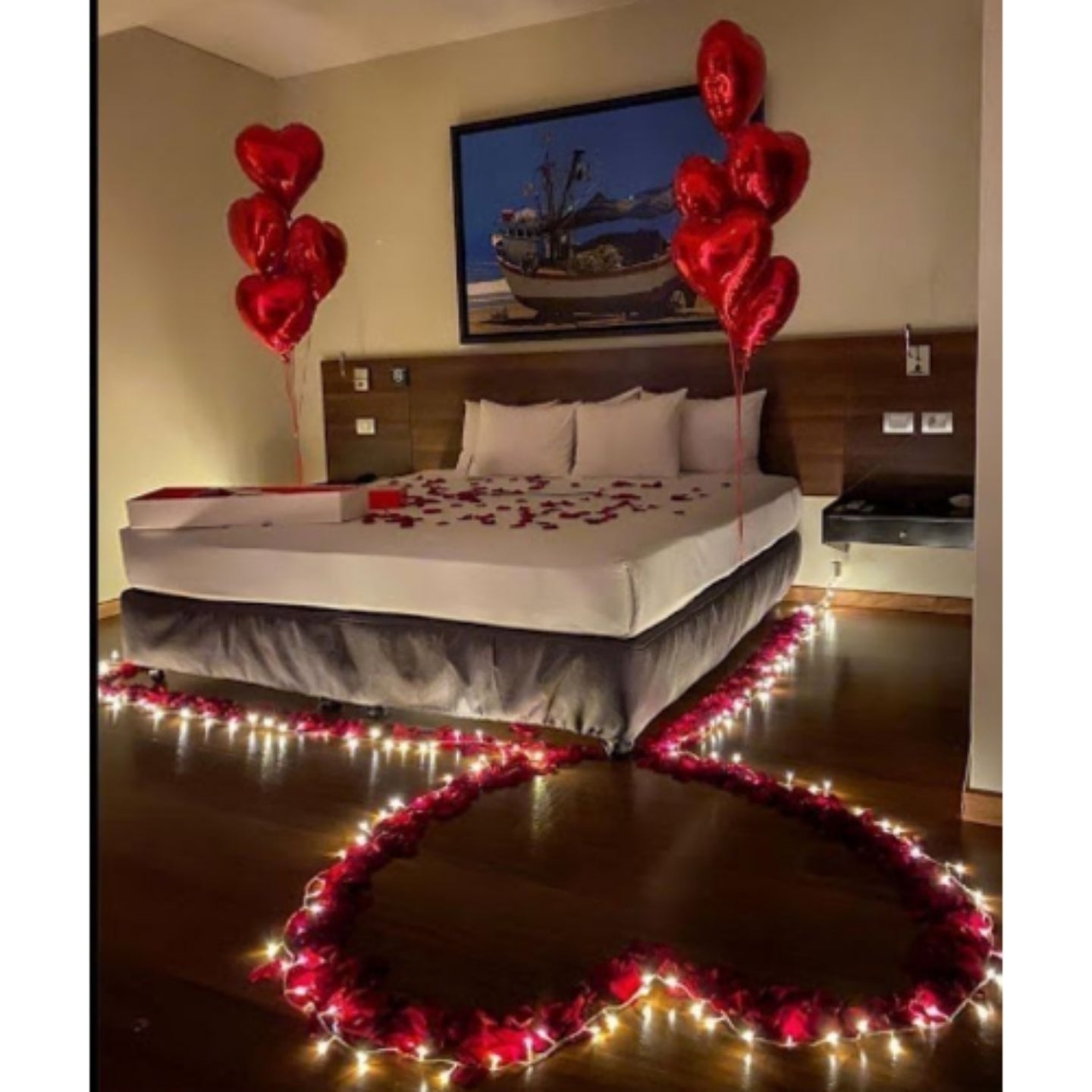 Decorated bedroom with heart-shaped balloons, rose petals, and fairy lights on a bed.