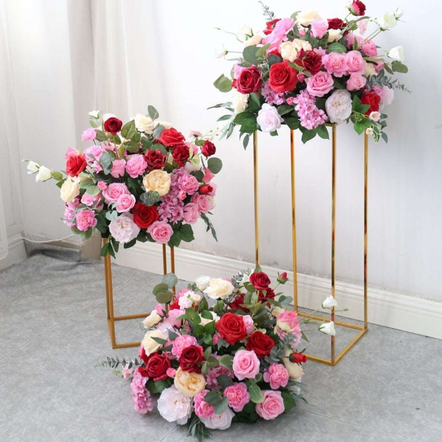 Three floral arrangements on gold stands against a white wall.