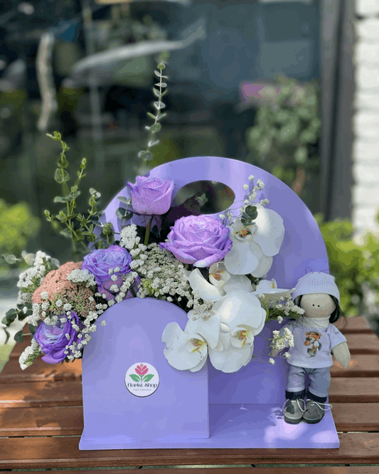 Floral arrangement with purple and white flowers on a purple stand with a small doll, set against a blurred outdoor background.