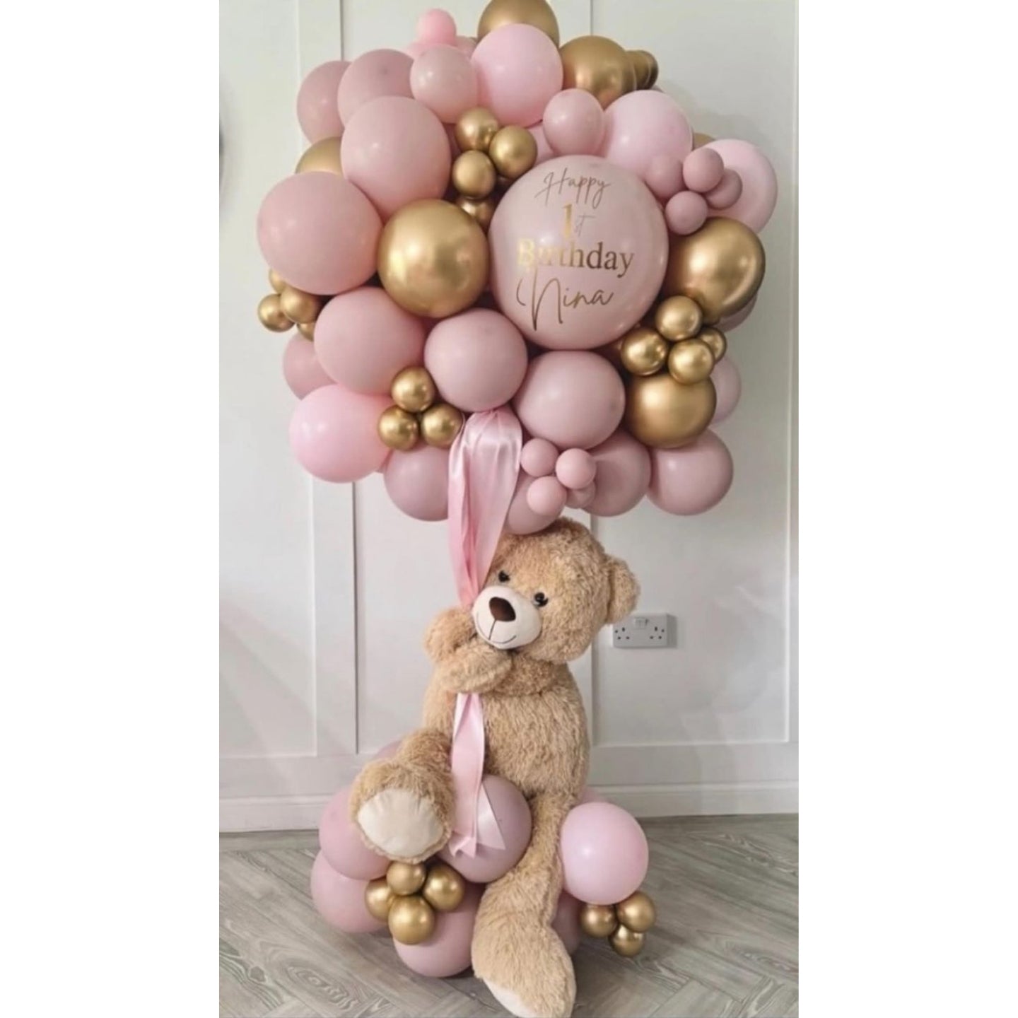 Balloon arrangement with a teddy bear and 'Happy Birthday Nina' balloon against a white wall.