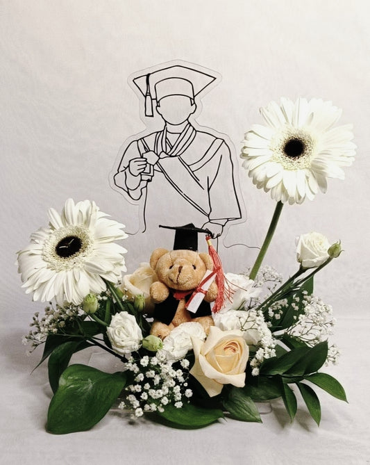 Graduation-themed arrangement with flowers, a teddy bear, and a cutout of a graduate on a white background.