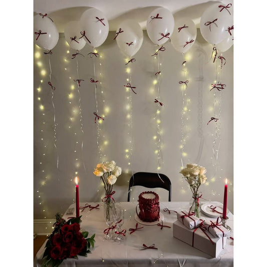 Decorated table with balloons, string lights, candles, and flowers against a wall.