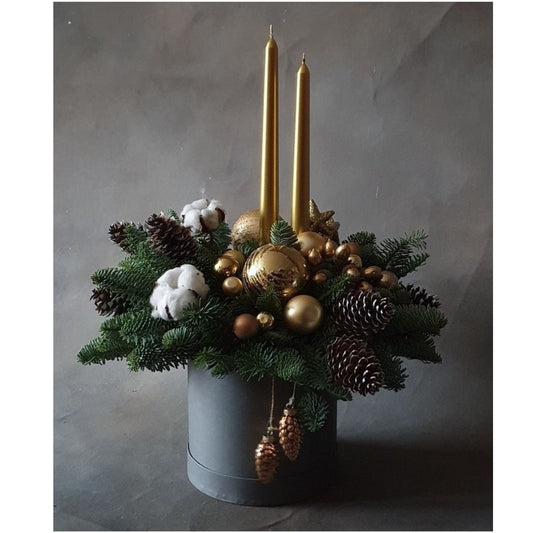 Decorative arrangement with gold candles, ornaments, and greenery in a gray container on a gray background