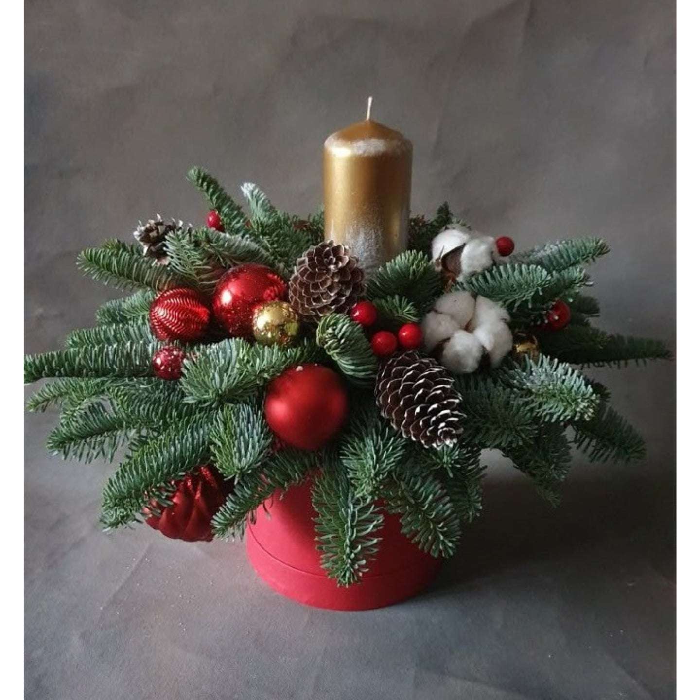 Decorative Christmas arrangement with a gold candle, red ornaments, and greenery on a gray background