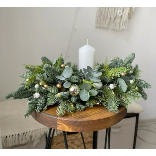 Decorative table centerpiece with greenery, small gold balls, and a white candle on a wooden table.
