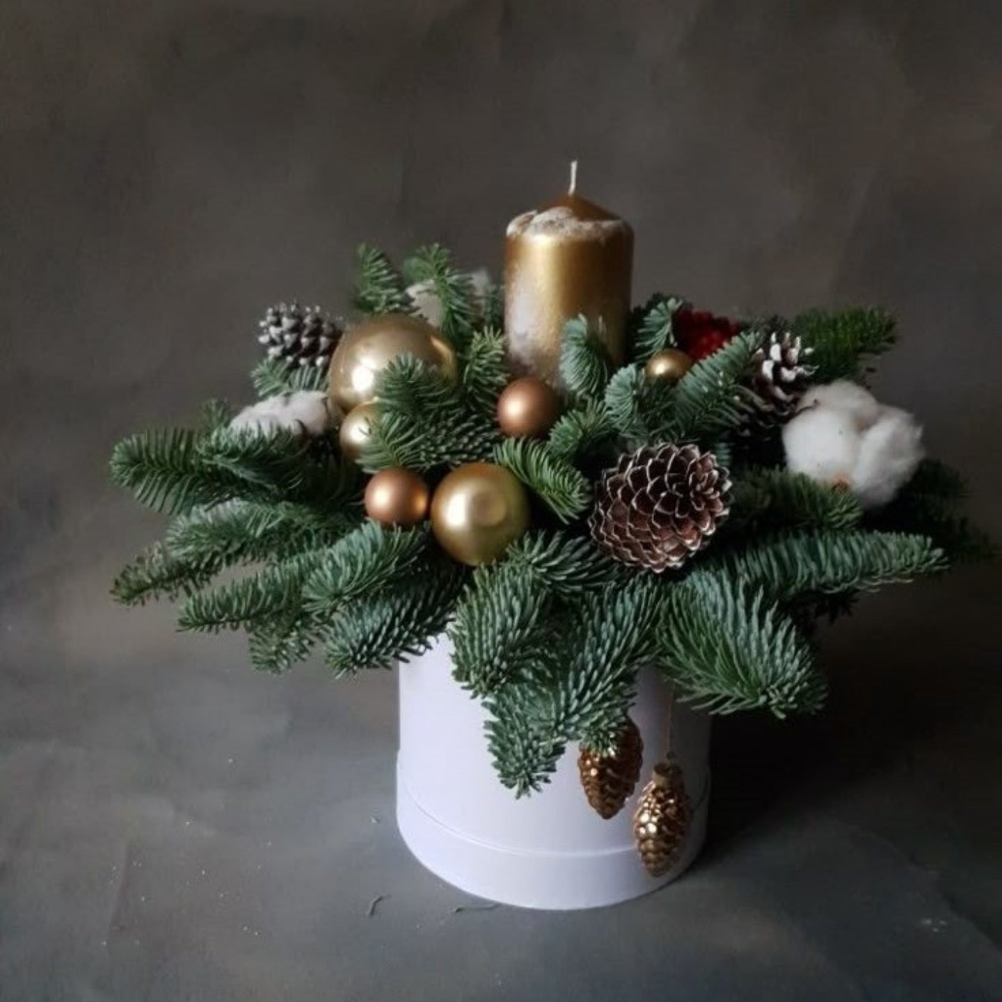 Decorative arrangement with greenery, gold balls, pinecones, and a candle in a white pot on a dark background