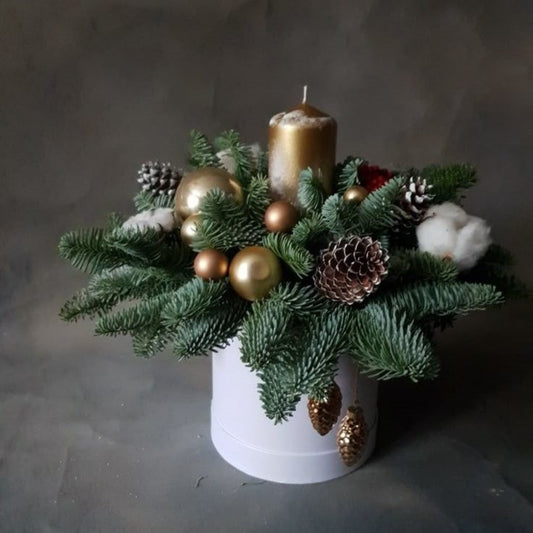 Decorative arrangement with greenery, gold balls, pinecones, and a candle in a white pot on a dark background