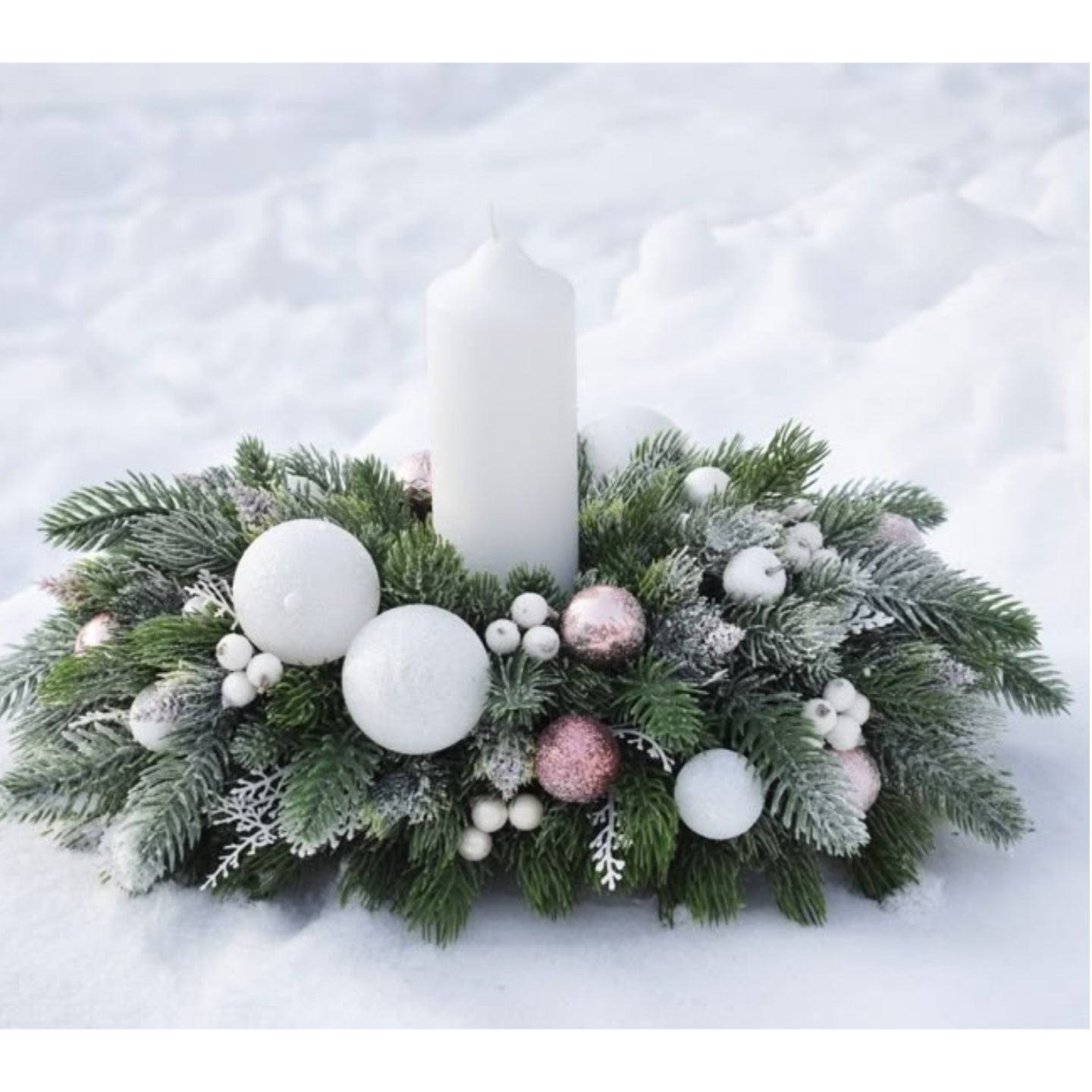Decorative Christmas arrangement with a candle surrounded by greenery, ornaments, and white snow on a snowy background.