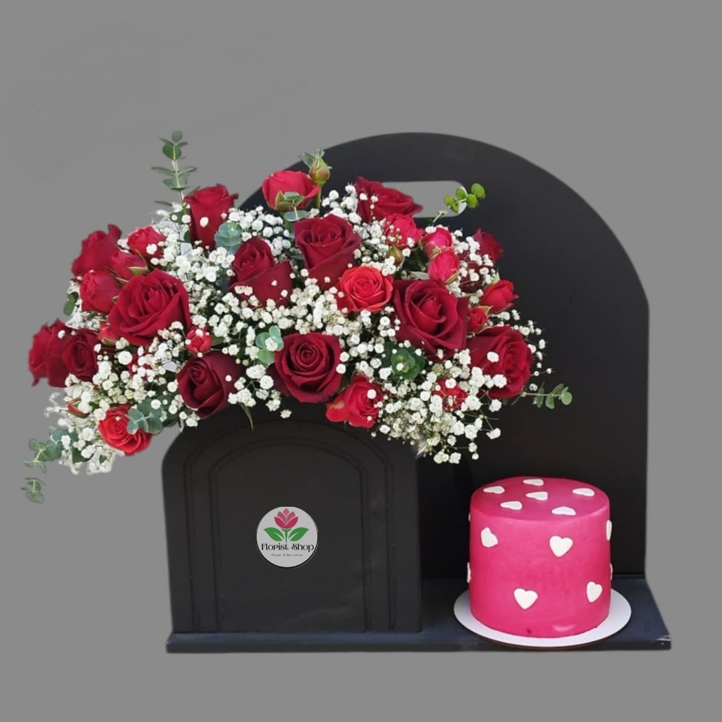 Black urn with red roses and white flowers, accompanied by a pink cake with heart patterns on a gray background.