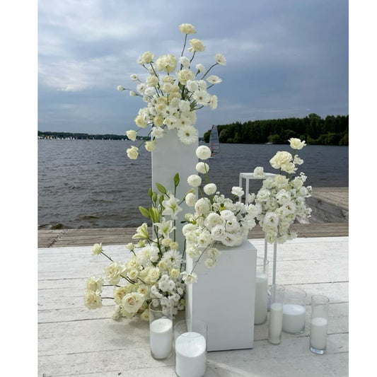 White floral arrangements and candles on a wooden deck by a body of water.
