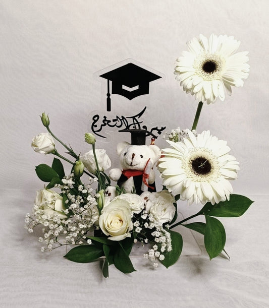Floral arrangement with a teddy bear and graduation cap on a light gray background