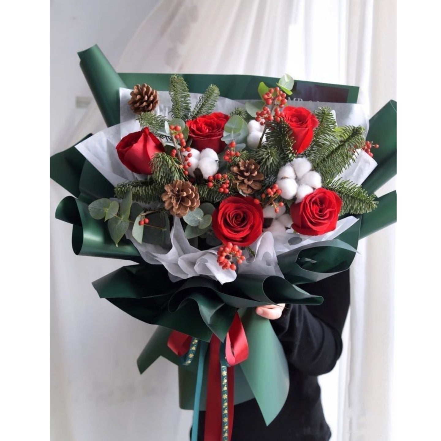 Bouquet of red roses with greenery and decorative ribbons held by a person.