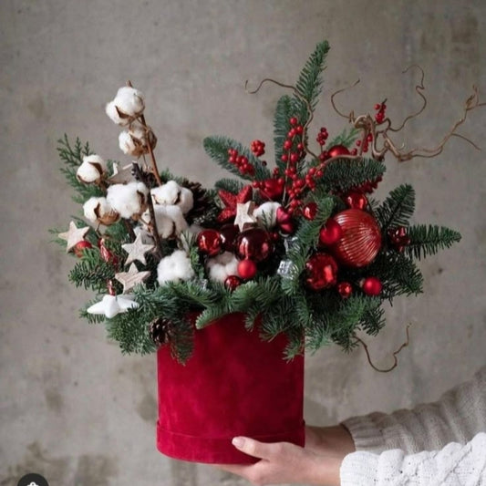 Decorative Christmas arrangement with red ornaments, cotton, and greenery in a red container against a neutral background.