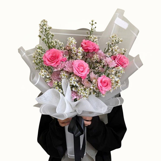 Bouquet of pink roses and white flowers wrapped in clear plastic with a black ribbon, held by a person wearing a black outfit.