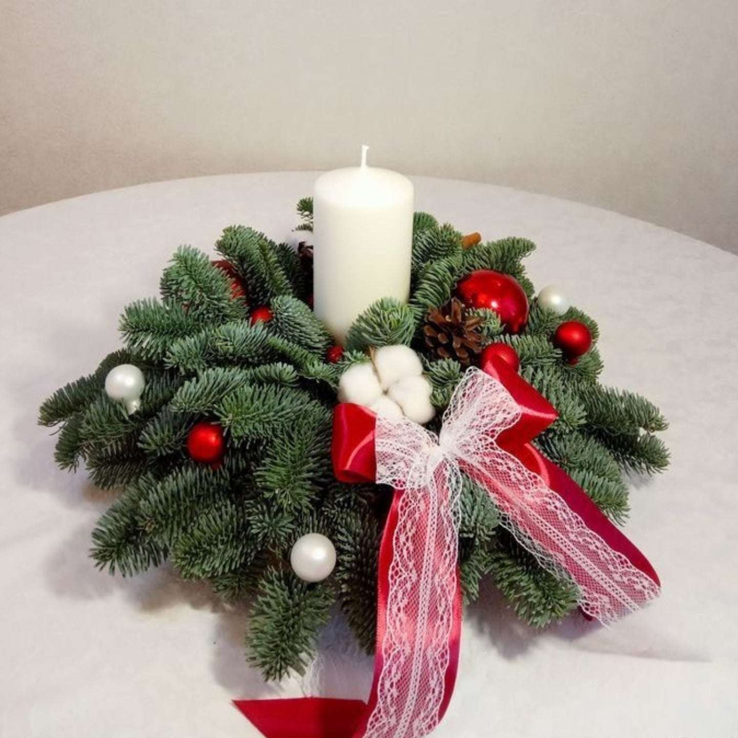 Christmas wreath with a candle, red and white ornaments, and a red ribbon on a white surface.