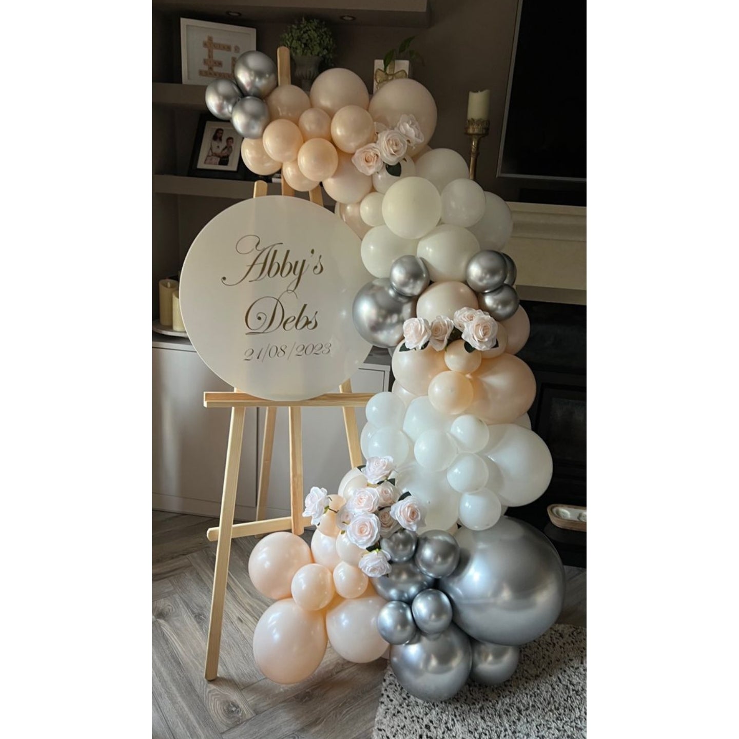 White, Pink and Silver Balloon arch with flowers and a sign on an easel in a room setting.