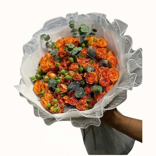 Bouquet of orange roses with greenery wrapped in white paper held by a person.