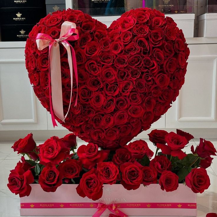 Heart-shaped arrangement of red roses with a pink ribbon on a white surface.